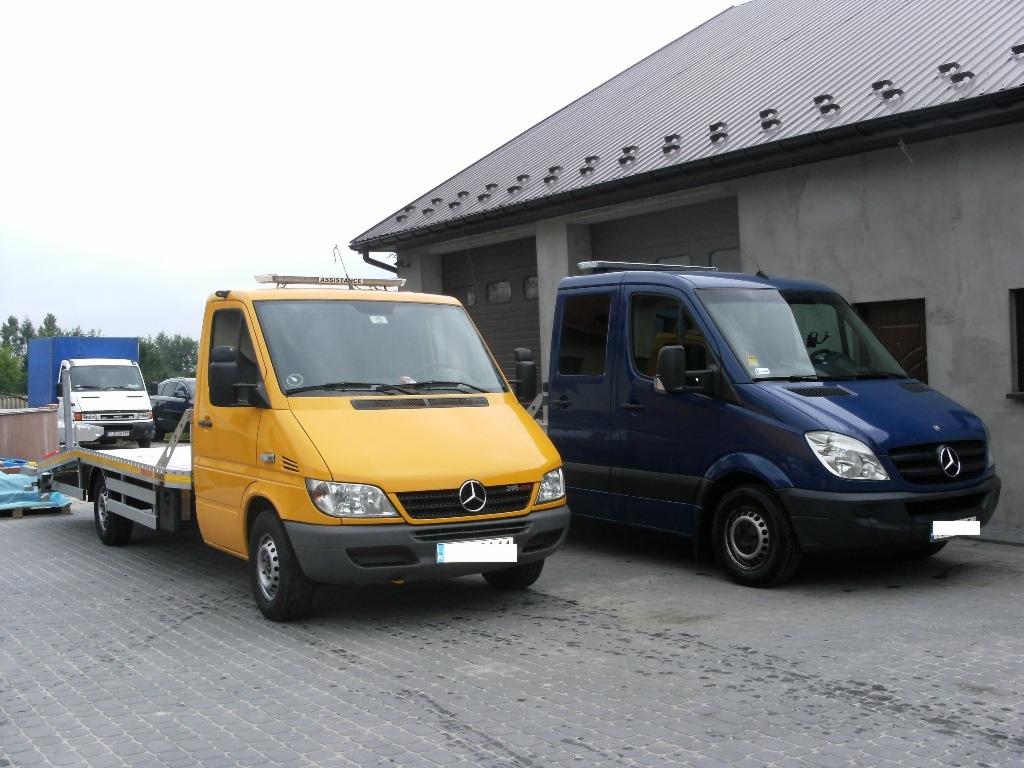 Żółta autolaweta Mercedes-Benz Sprinter z platformą i niebieski bus Mercedes-Benz Sprinter na szarym brukowanym placu przed budynkiem z blachy trapezowej.