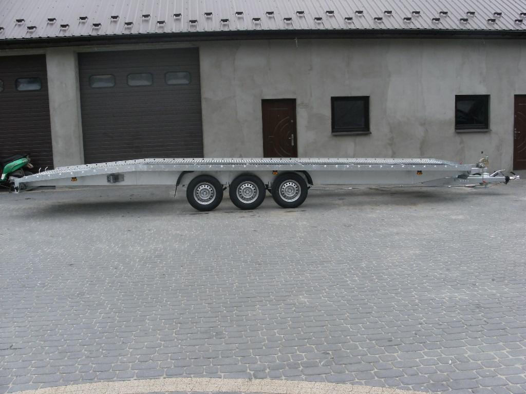 Długa, srebrna laweta z trzema osiami, zaparkowana na brukowanym placu przed budynkiem z szarymi drzwiami garażowymi i oknami.