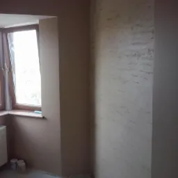 Ściana z efektem dekoracyjnym w odcieniach beżu, widok na okno z drewnianą ramą, fragment grzejnika i narzędzia malarskie na podłodze. Wykończenie wnętrza.