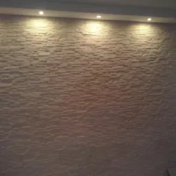 Ściana z dekoracyjnej, beżowej cegły, oświetlona od góry punktowymi lampami. Widoczna faktura i ułożenie cegiełek, nadające wnętrzu nowoczesny wygląd.