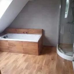 Łazienka na poddaszu z drewnianą obudową wanny, kabiną prysznicową i drewnianą podłogą. Ściany wykończone szarą fakturą, okno dachowe nad wanną.