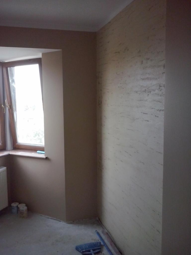 Ściana z efektem dekoracyjnym w odcieniach beżu, widok na okno z drewnianą ramą, fragment grzejnika i narzędzia malarskie na podłodze. Wykończenie wnętrza.