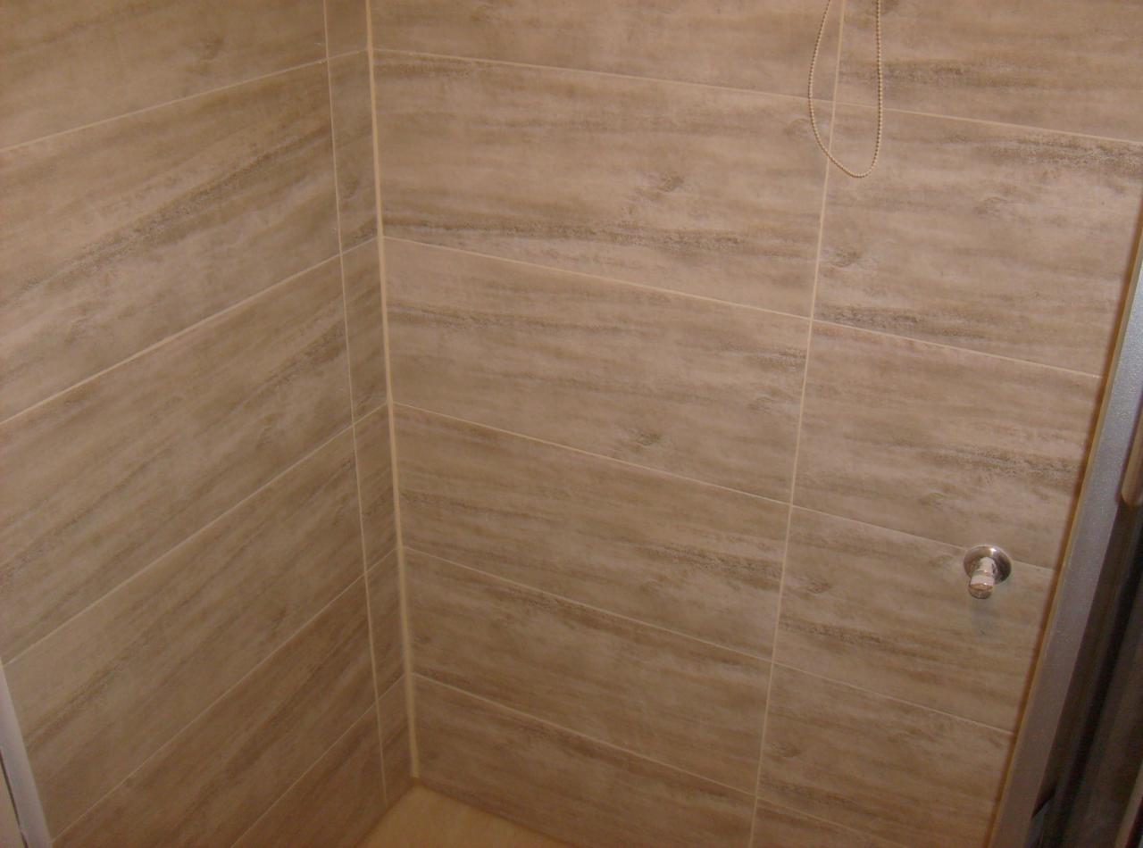 Beżowe, poziome płytki imitujące drewno w kabinie prysznicowej. Widoczna bateria i fragment łańcuszka. Minimalistyczny design, ciepłe odcienie.