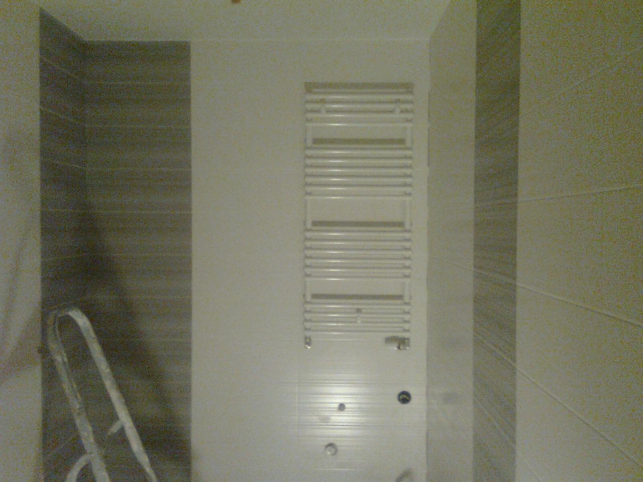 Wykończona łazienka z grzejnikiem drabinkowym na tle płytek w odcieniach szarości i beżu. Fragment drabiny malarskiej w lewym dolnym rogu.