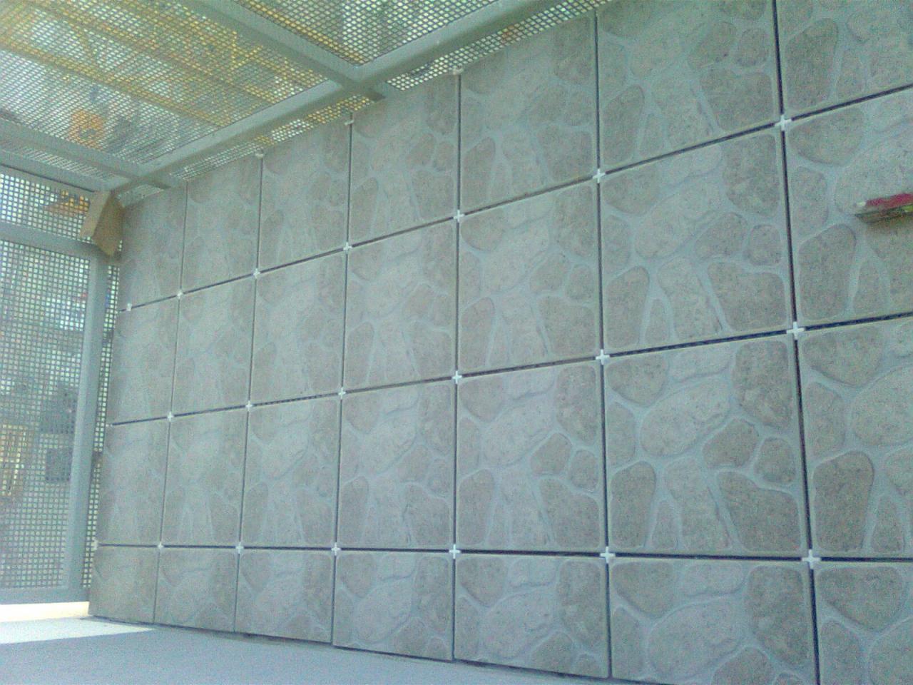 Ściana wyłożona beżowymi płytkami ceramicznymi o nieregularnym wzorze, z krzyżykami dystansowymi, widoczna z dołu pod lekkim kątem, przy kratownicy.