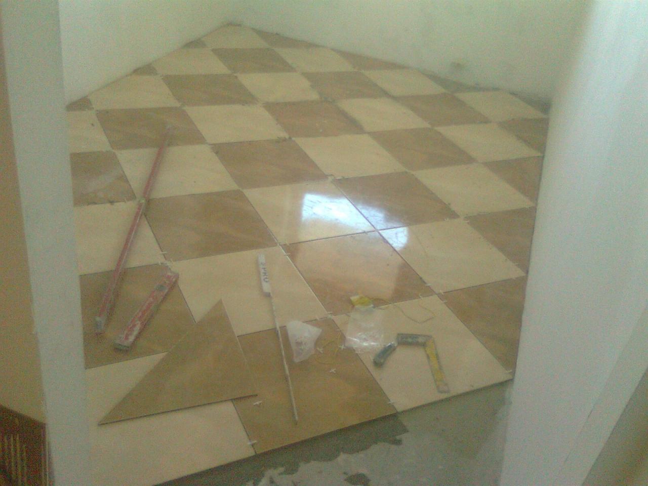 Układanie podłogi z płytek w szachownicę; widoczne narzędzia glazurnicze i fragment ściany. Praca w toku, płytki w odcieniach beżu i brązu.