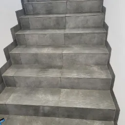 Szare, ceramiczne schody wyłożone płytkami z delikatnym wzorem, przylegające do białej ściany, z niebieską szpachelką widoczną na dole po lewej stronie.