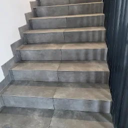 Schody wyłożone szarymi płytkami ceramicznymi z delikatnym wzorem, widoczne z dołu, przy ścianie i metalowej balustradzie, z fragmentem mopa w rogu.