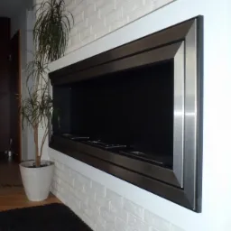 Wnęka kominkowa z metalowym obramowaniem, biała ceglana ściana, roślina w doniczce i czarny dywanik w nowoczesnym wnętrzu.