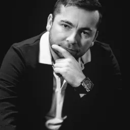 Czarno-białe portretowe ujęcie mężczyzny w garniturze, z brodą i zegarkiem na ręce, zamyślonego, z palcem przy brodzie, na ciemnym tle.