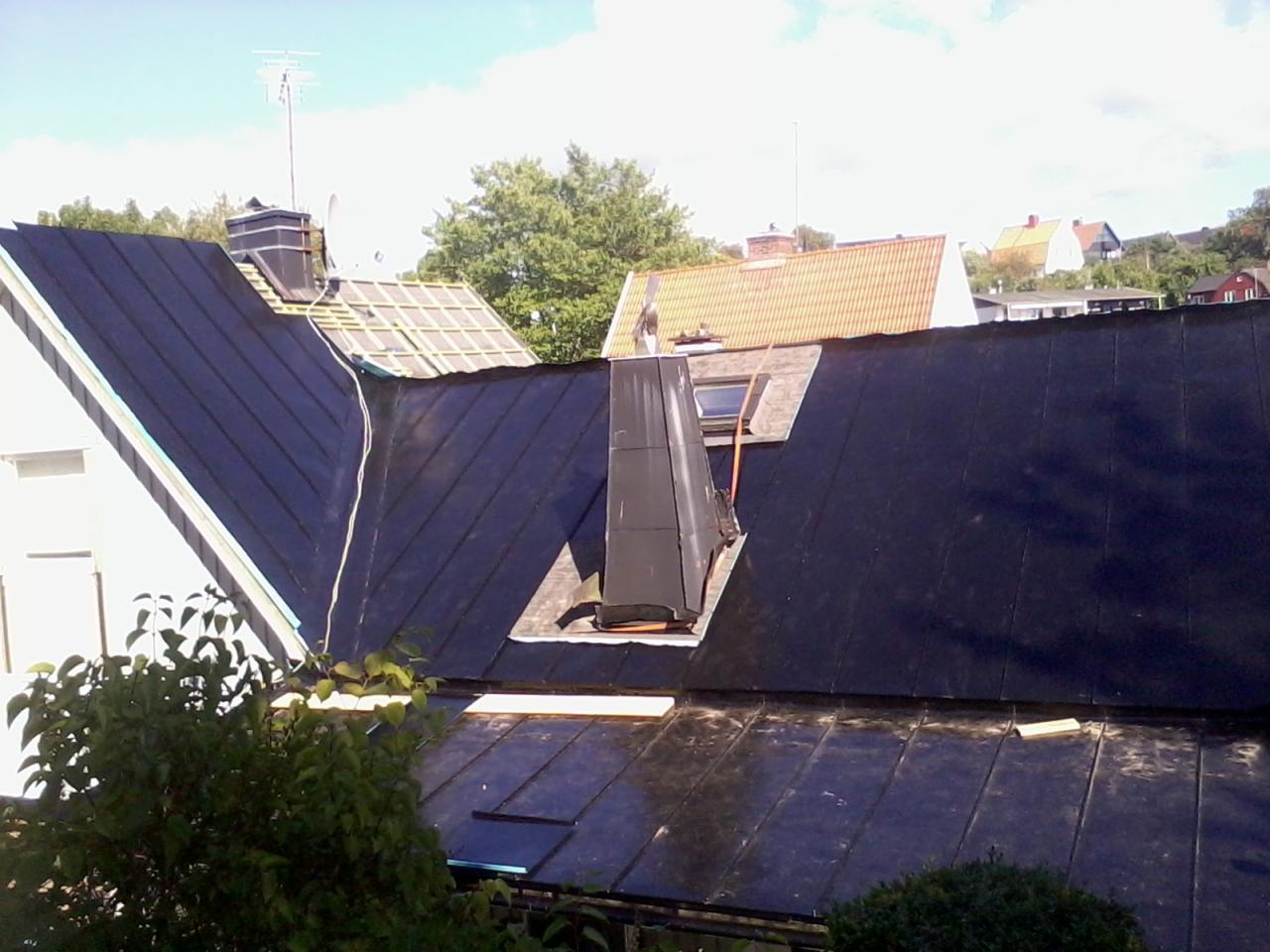 Widok z góry na dach z czarną papą, fragment dachu z drewnianą konstrukcją, komin z metalowym obiciem i fragmenty innych budynków w tle.