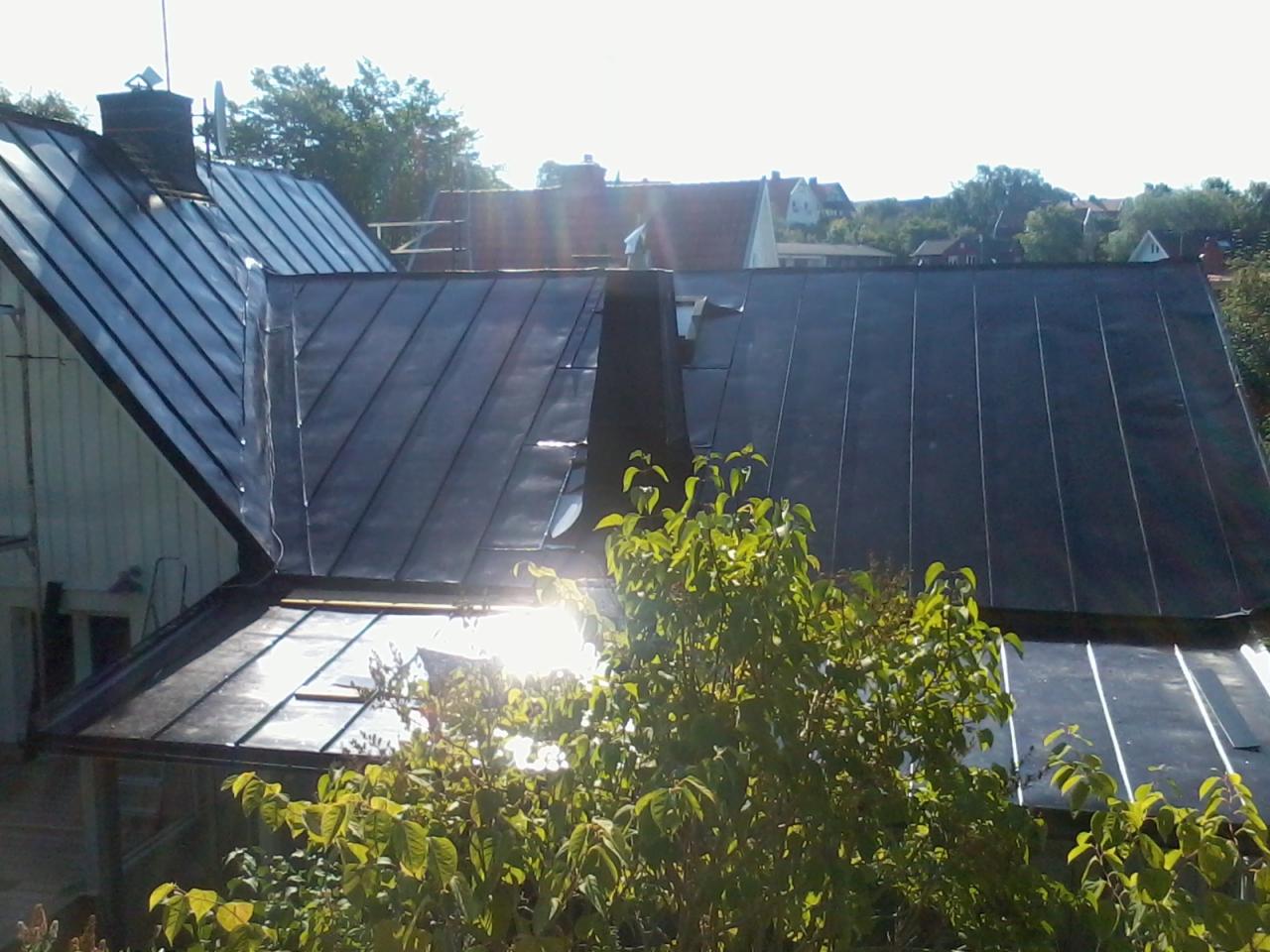 Ciemna, metalowa dachówka na domu jednorodzinnym, widziana z perspektywy ogrodu, z kominem i anteną na tle zieleni i innych budynków w oddali.