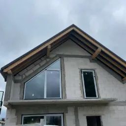 Dom w trakcie budowy z zamontowanymi oknami o różnych kształtach, w tym trójkątnym na poddaszu. Widoczny fragment samochodu dostawczego.