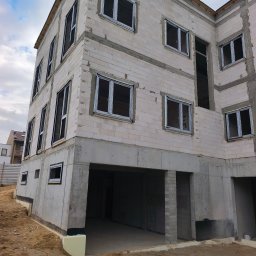 Producent Okien PCV i ALU Patryk Pankowski - Surowy stan budynku w trakcie montażu stolarki okiennej. Widoczne otwory okienne z zamontowanymi ramami. Budynek w trakcie budowy, ściany z betonu komórkowego.