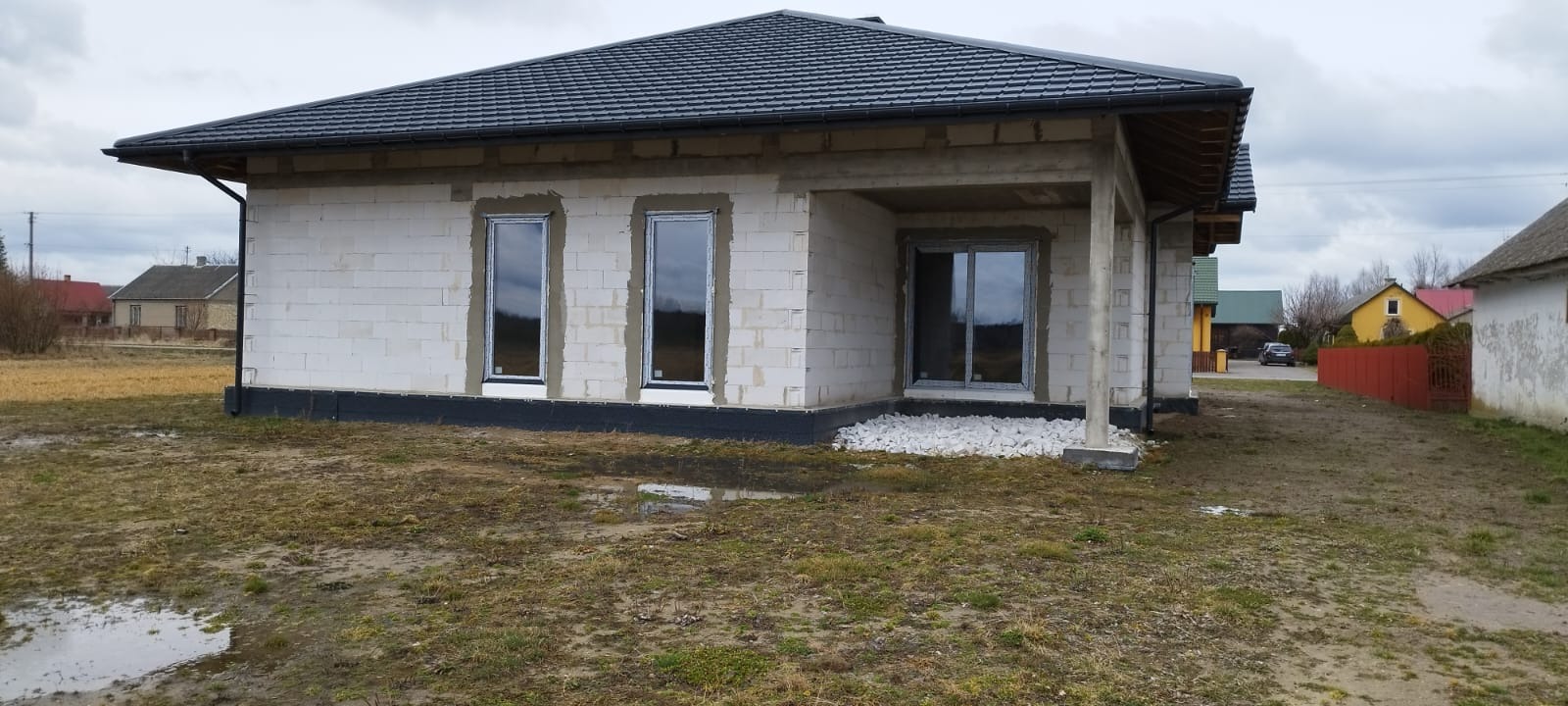 Surowy stan nowego domu z zamontowanymi oknami z białymi ramami, widoczne białe pustaki, czarna hydroizolacja fundamentów i szara dachówka.