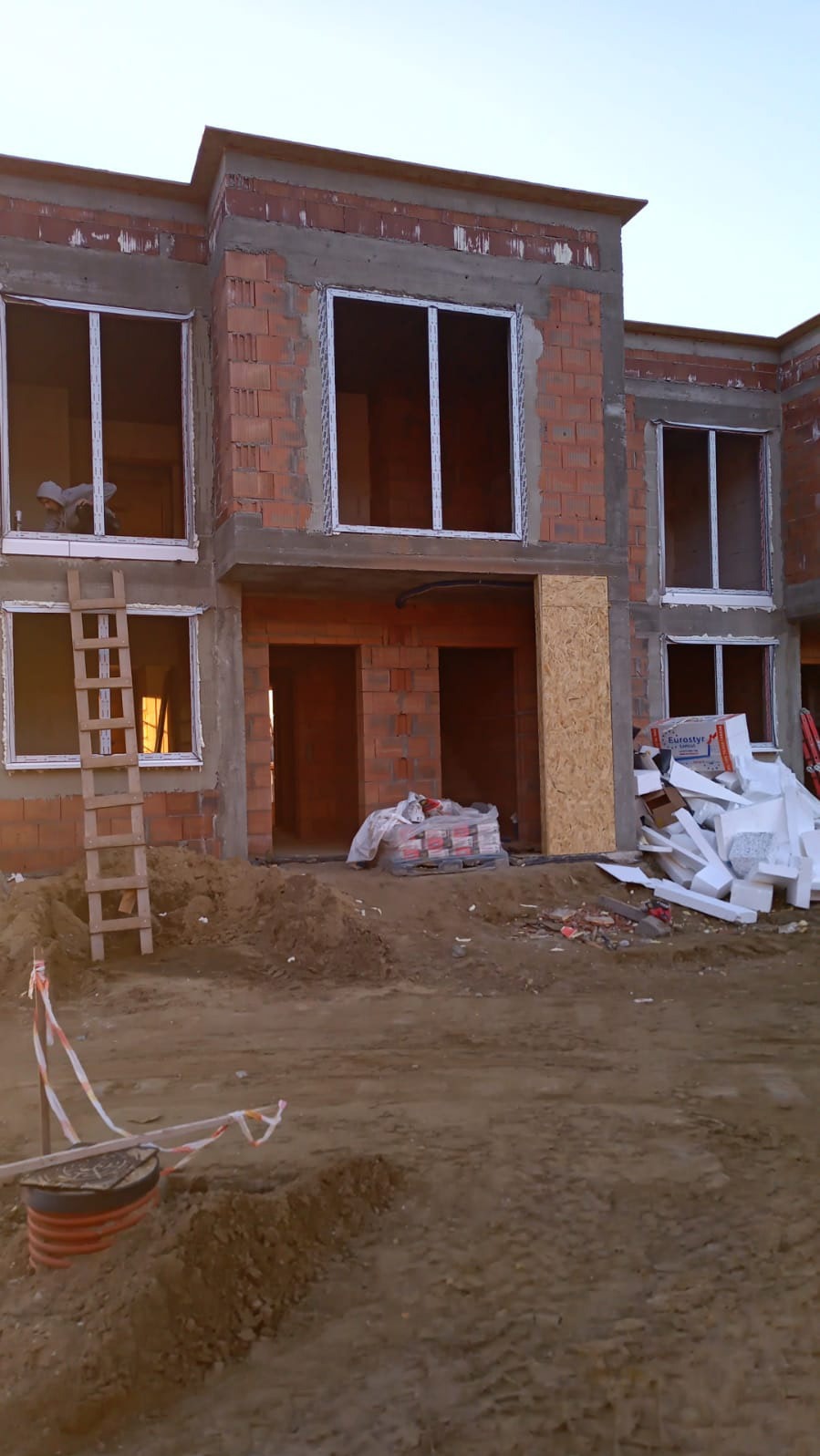 Surowy stan budynku w trakcie montażu stolarki okiennej. Widoczne białe ramy okienne w murach z cegły, drabina oparta o ścianę, materiały budowlane na ziemi.