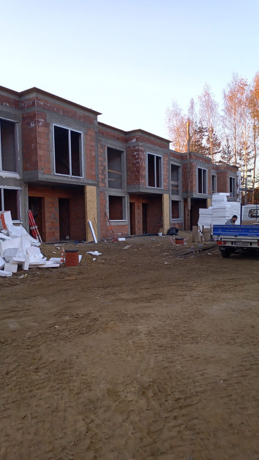Surowy stan budowy szeregowców z pustymi otworami okiennymi, widoczne materiały izolacyjne i ciężarówka z napisem 'Montaż okien, bram, rolet zewnętrznych'.