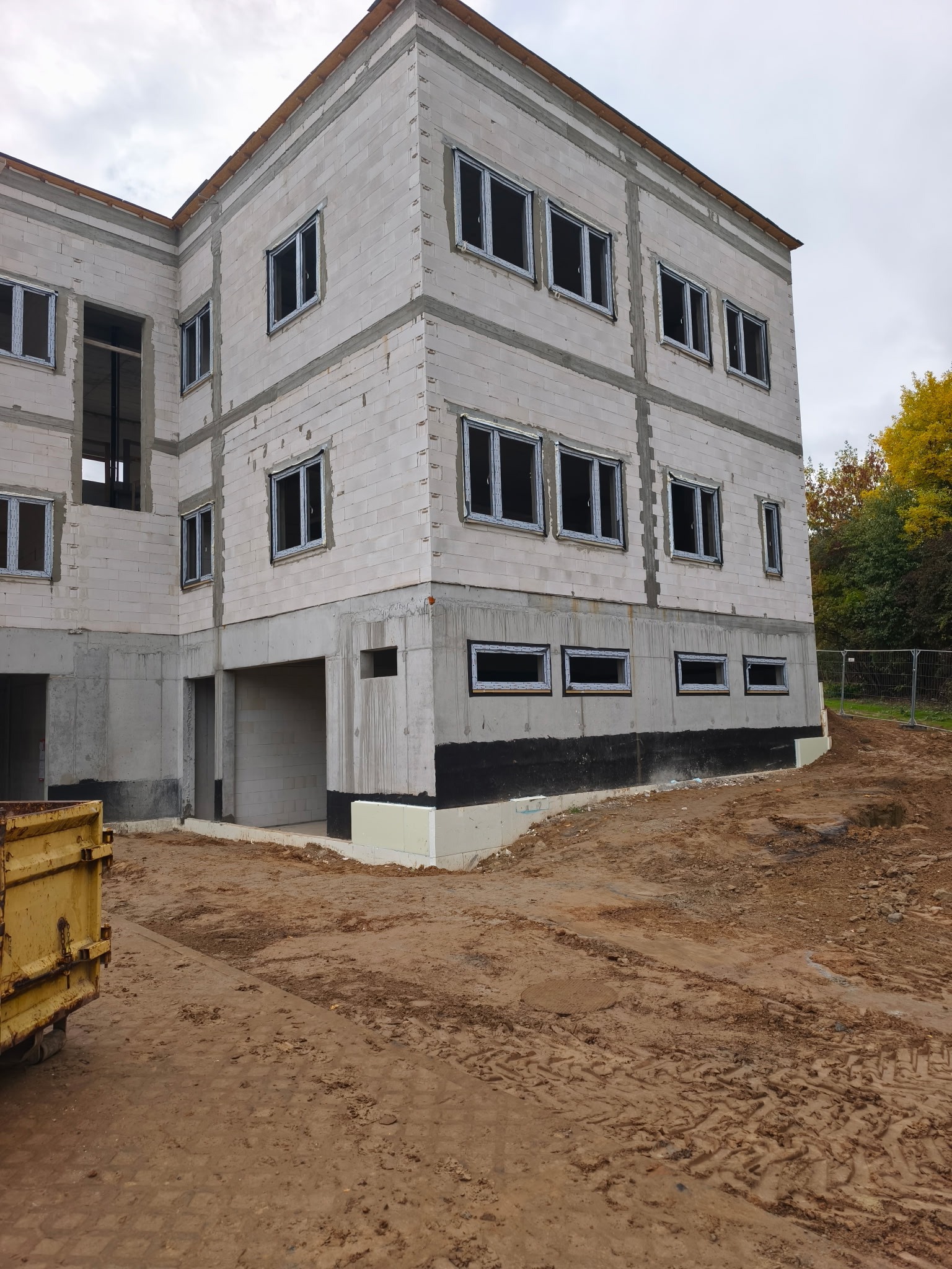 Budynek w stanie surowym otwartym z zamontowanymi oknami, widoczne bloczki betonowe i teren budowy. Częściowo ocieplony fundament.
