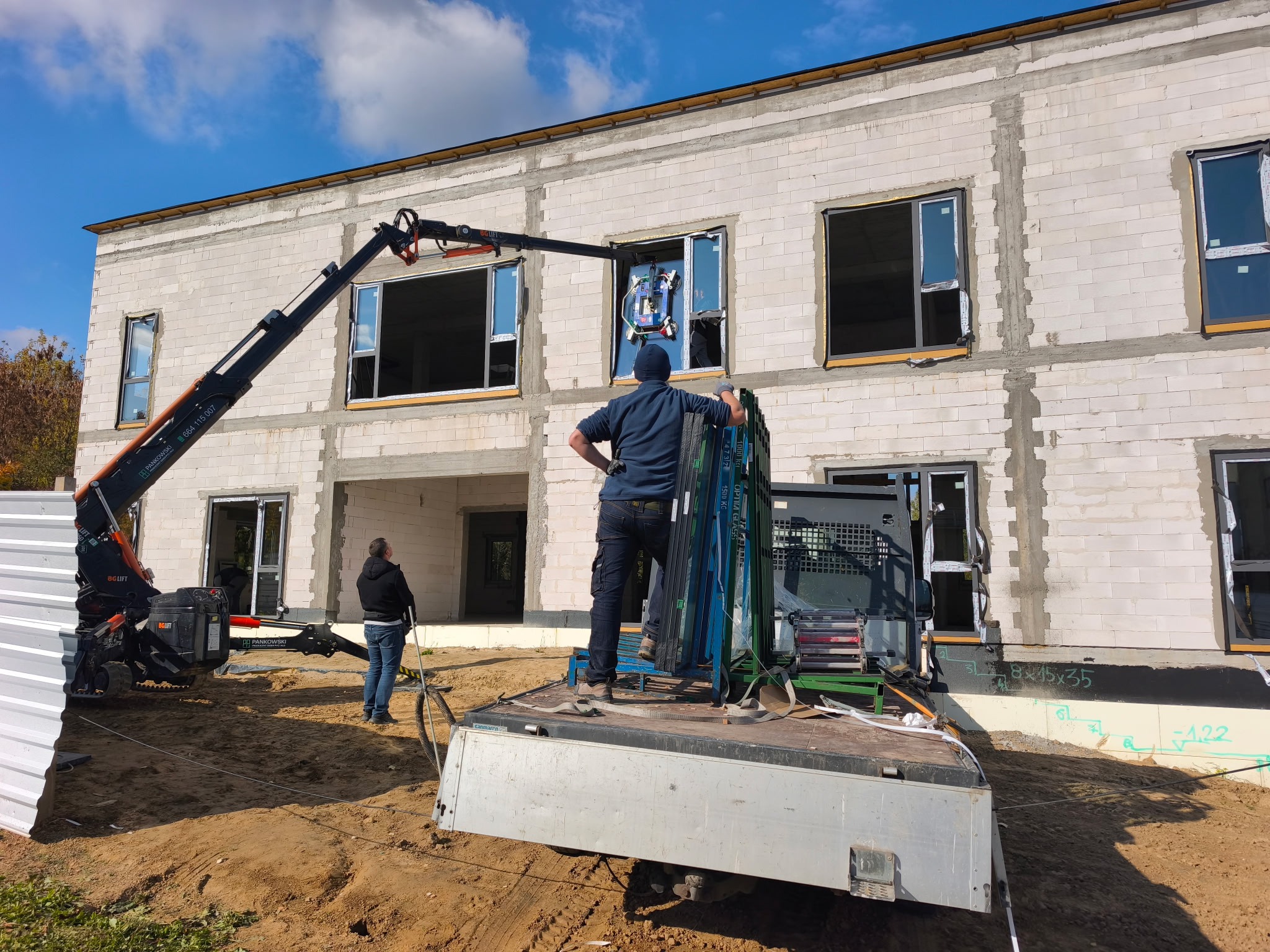 Montaż okna na budowie z użyciem dźwigu ssącego. Widoczny budynek w trakcie budowy, pracownicy i okno w trakcie instalacji. Jasne niebo w tle.