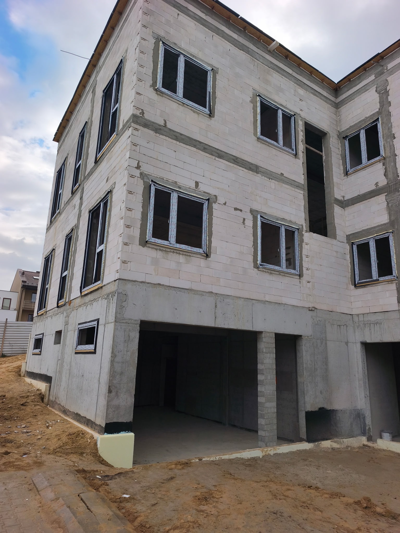 Surowy stan budynku w trakcie montażu stolarki okiennej. Widoczne otwory okienne z zamontowanymi ramami. Budynek w trakcie budowy, ściany z betonu komórkowego.