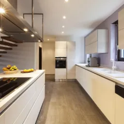 Nowoczesna kuchnia z wyspą, białe fronty szafek, płyta indukcyjna, drewniane schody w tle, oświetlenie punktowe i minimalistyczny design.