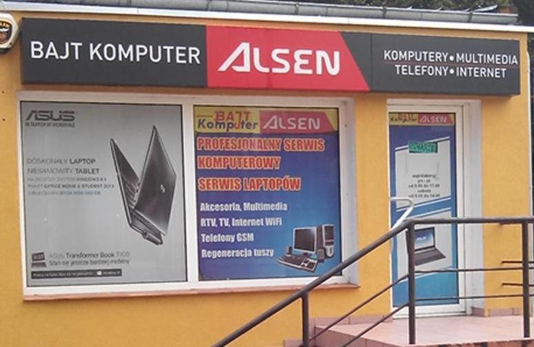 Fasada sklepu komputerowego 'Bajt Komputer Alsen' z reklamami laptopów i usług serwisowych, w tym regeneracji tuszy i akcesoriów. Widoczny szyld i okna wystawowe.