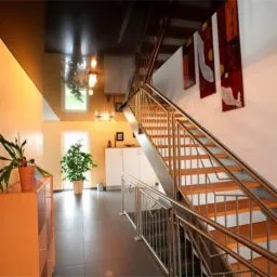 Jasne wnętrze z ciemnym, lustrzanym sufitem, na którym odbijają się lampy. Widoczne schody z metalową balustradą i rośliny doniczkowe.