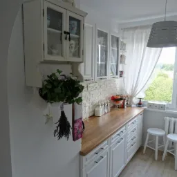 Jasna kuchnia w stylu rustykalnym z białymi szafkami, drewnianym blatem, ceglaną ścianą i oknem z białymi zasłonami, uzupełniona o roślinę w doniczce i bukiet lawendy.