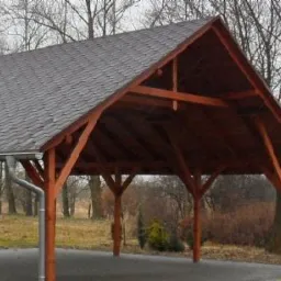 Zadaszenie z drewnianą konstrukcją i pokryciem z gontu bitumicznego. Widoczne rynny i słupy nośne. Tło stanowią drzewa i krzewy w jesiennej scenerii.