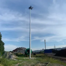 Wysoki maszt oświetleniowy z wieloma reflektorami, stojący na terenie przemysłowym z hałdami złomu i torami kolejowymi w tle, widok w słoneczny dzień.
