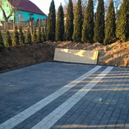 Częściowo ułożona, grafitowa kostka brukowa z białymi pasami na podjeździe, widok z góry. W tle ziemia, tuje i dom.