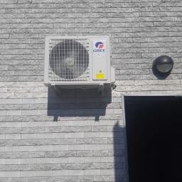 CST Centrum Serwisu Technicznego Paweł Redel - Zewnętrzna jednostka klimatyzatora marki Gree zamontowana na elewacji z szarej cegły, widoczne ostrzeżenie na obudowie i lampa zewnętrzna obok.