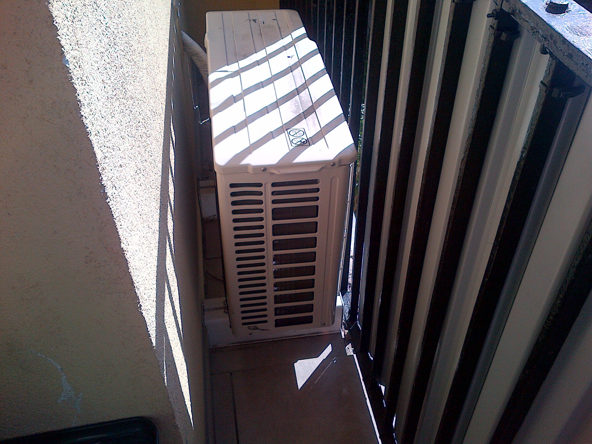 Zewnętrzna jednostka klimatyzacji zamontowana na balkonie, widoczne żaluzje i cień rzucany przez nie na urządzenie.