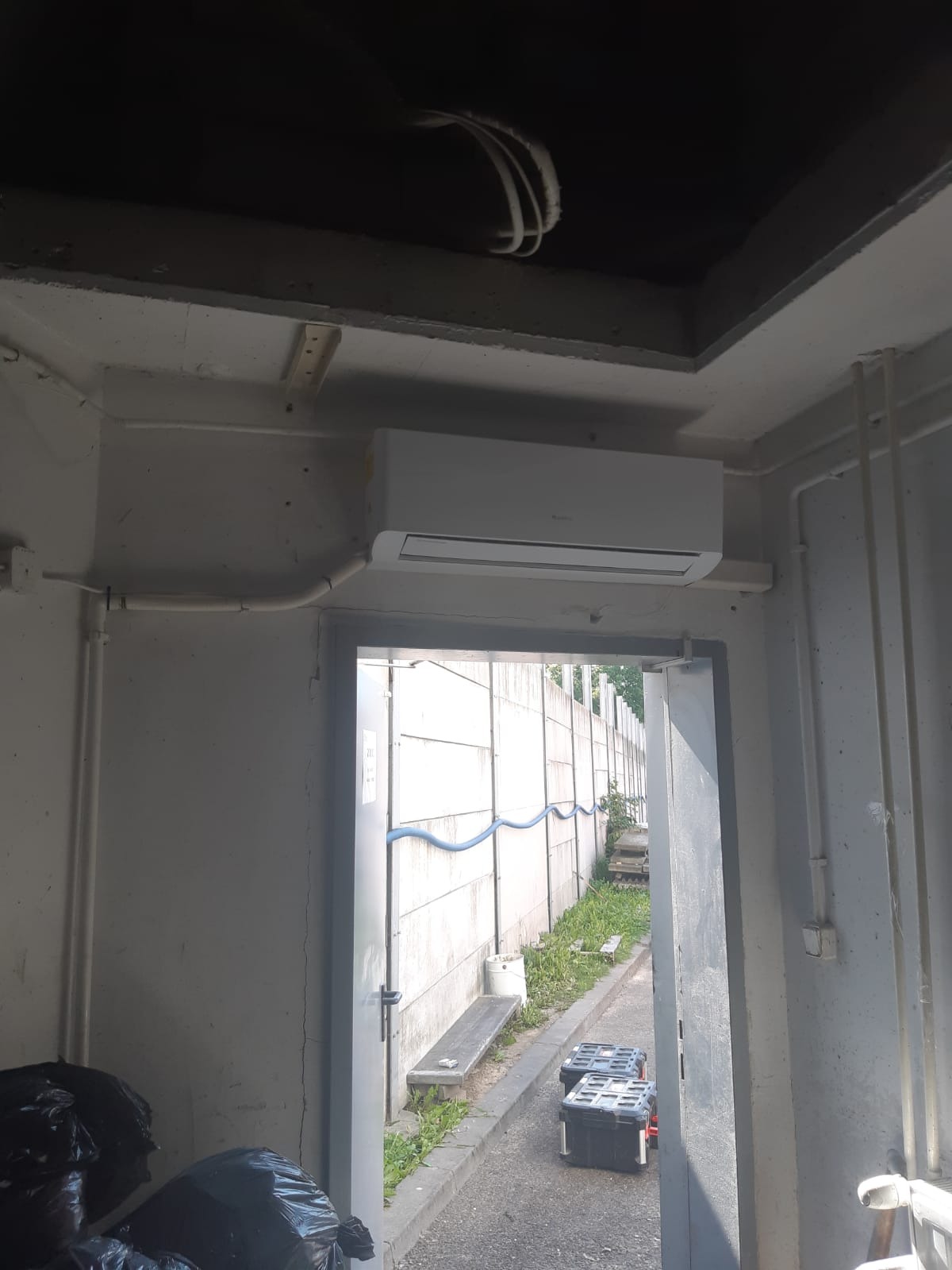 Wnętrze pomieszczenia z zamontowanym klimatyzatorem na ścianie, widoczne rury instalacyjne i skrzynki narzędziowe na zewnątrz