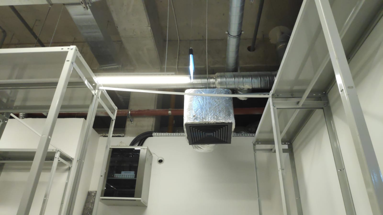 Kwadratowy wylot powietrza z instalacji, owinięty folią aluminiową, zawieszony pod sufitem w pomieszczeniu z widocznymi rurami i przewodami, oświetlony jarzeniówką.