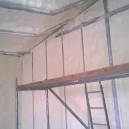 Ściany i sufit budynku pokryte jasną pianą izolacyjną, widoczne metalowe profile konstrukcyjne i fragment rusztowania z drewnianą deską na pierwszym planie.