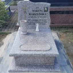 Szary, granitowy nagrobek z krzyżem i wizerunkiem zmarłej, z inskrypcją zawierającą daty urodzin i śmierci, ustawiony na cmentarzu.
