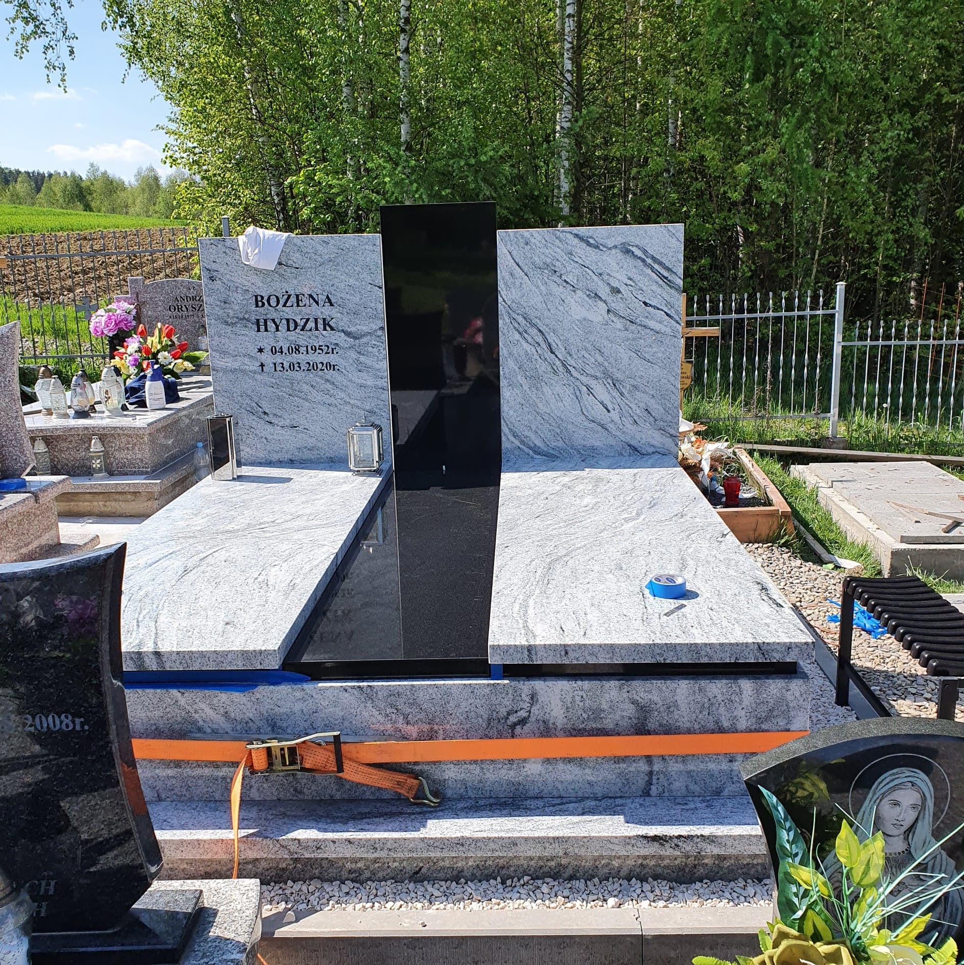 Nowo postawiony, jasnoszary nagrobek z czarnym elementem centralnym, zabezpieczony pomarańczowym pasem transportowym i niebieską taśmą, na cmentarzu z innymi nagrobkami i zielenią w tle.