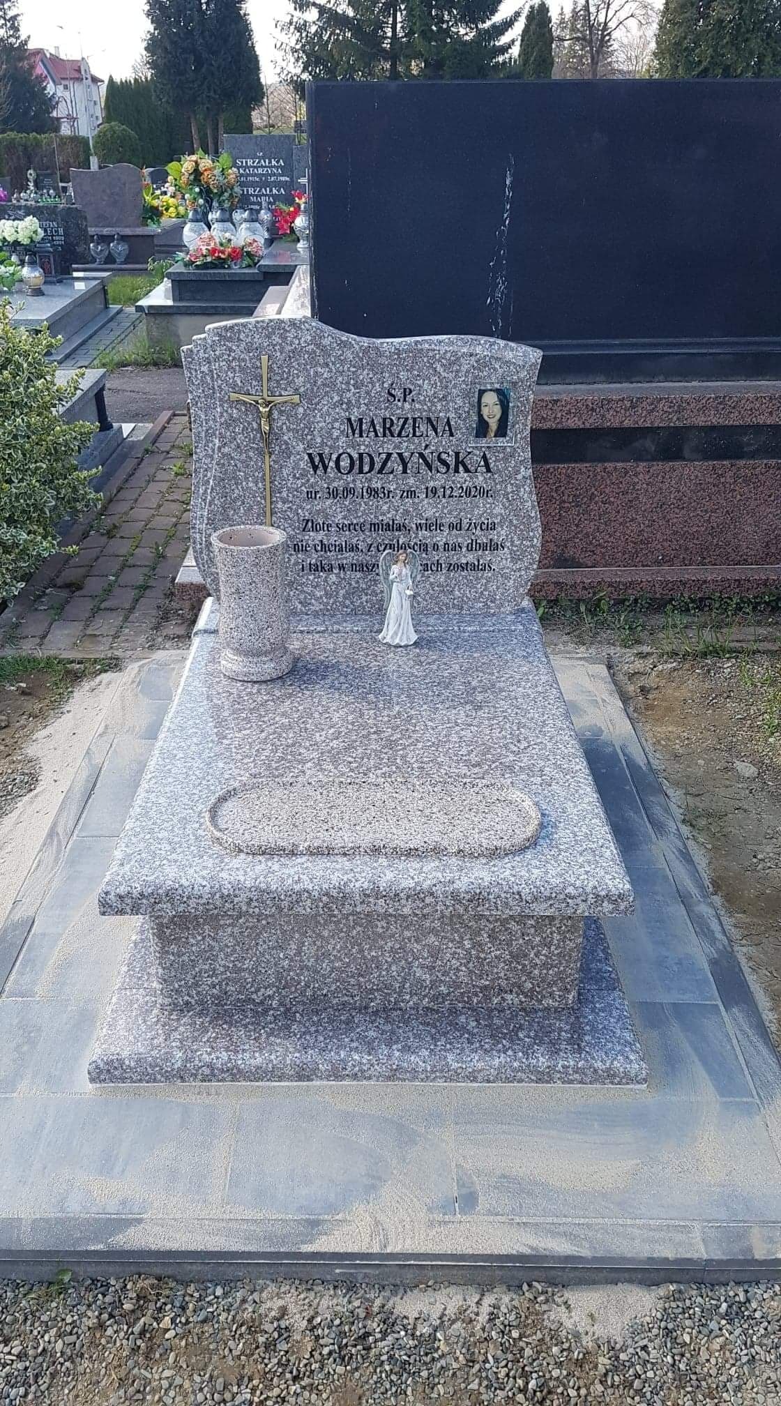 Szary, granitowy nagrobek z krzyżem i wizerunkiem zmarłej, z inskrypcją zawierającą daty urodzin i śmierci, ustawiony na cmentarzu.
