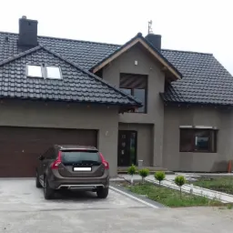 Nowoczesny dom jednorodzinny z ciemną dachówką, grafitową elewacją i brązową bramą garażową, na podjeździe stoi brązowy samochód Volvo, w tle łąka.