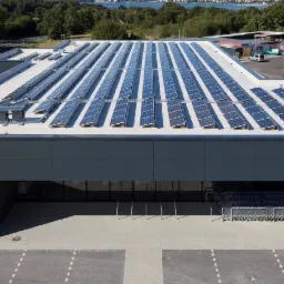 Widok z góry na budynek sklepu z panelami słonecznymi na dachu, logo sklepu widoczne na fasadzie, w tle drzewa i zbiornik wodny.