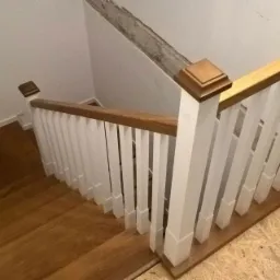 Widok z góry na drewniane schody z białymi balustradami i drewnianą poręczą, częściowo wykończone, z widoczną surową ścianą i podłogą w trakcie remontu.