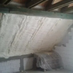 Poddasze w trakcie izolacji: ściana i skos dachu pokryte białą pianą izolacyjną, widoczne drewniane elementy konstrukcyjne i białe bloczki, materiały budowlane zabezpieczone folią.