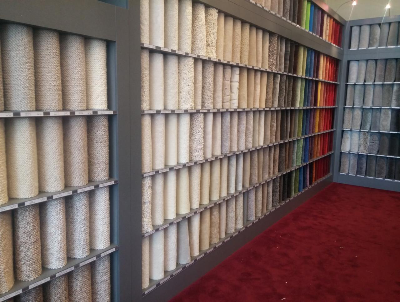 Ekspozycja próbek wykładzin w sklepie: rolki różnorodnych kolorów i faktur ułożone na półkach, widoczna czerwona wykładzina podłogowa.