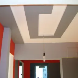 Wnętrze po malowaniu: pomarańczowa ściana, szara ściana i sufit z geometrycznym wzorem w odcieniach bieli i szarości. Widoczny fragment instalacji oświetleniowej.