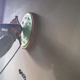Szlifowanie ściany szlifierką z tarczą, widoczne nierówności i ślady po obróbce. Praca w toku, jasne wnętrze, kabel zasilający.