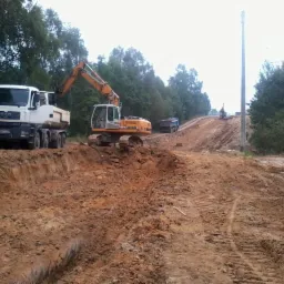 Żółta koparka Liebherr i biała ciężarówka MAN na placu budowy. W tle widać wykopany teren z drzewami i słupami energetycznymi. Prace ziemne w toku.