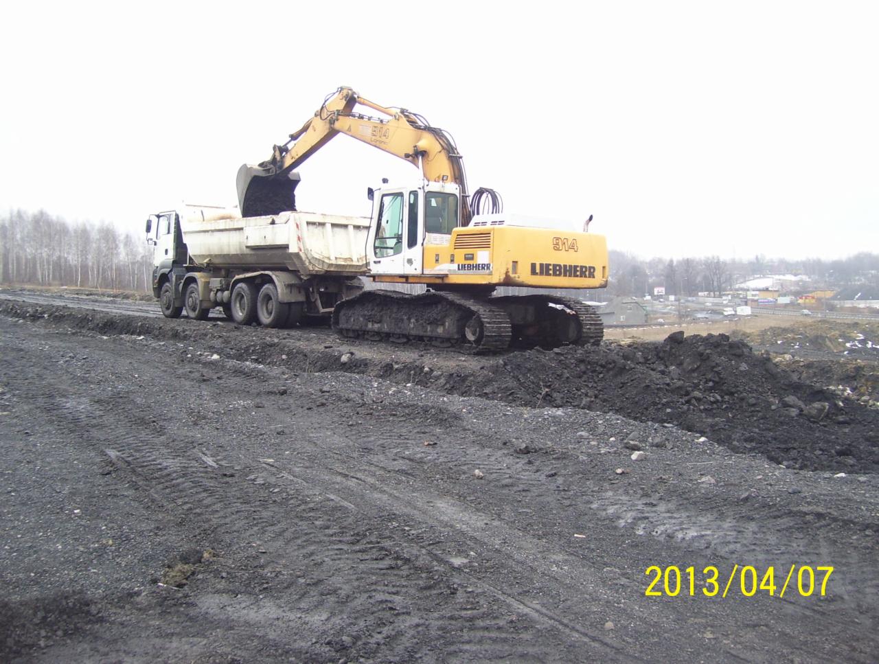 Żółto-biała koparka Liebherr 914 nabiera ziemię do białej ciężarówki na placu budowy. W tle widać drzewa i zabudowania. Ziemia ma wyraźne ślady opon.
