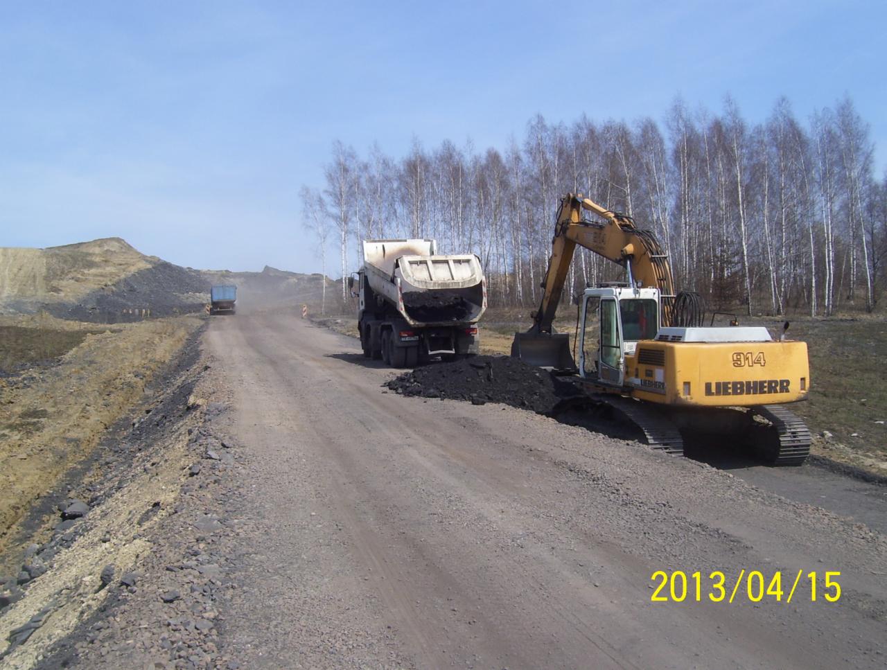 Żółta koparka Liebherr 914 ładuje urobek do wywrotki na szutrowej drodze. W tle widoczne drzewa i hałda ziemi. Ujęcie z 15 kwietnia 2013 roku.