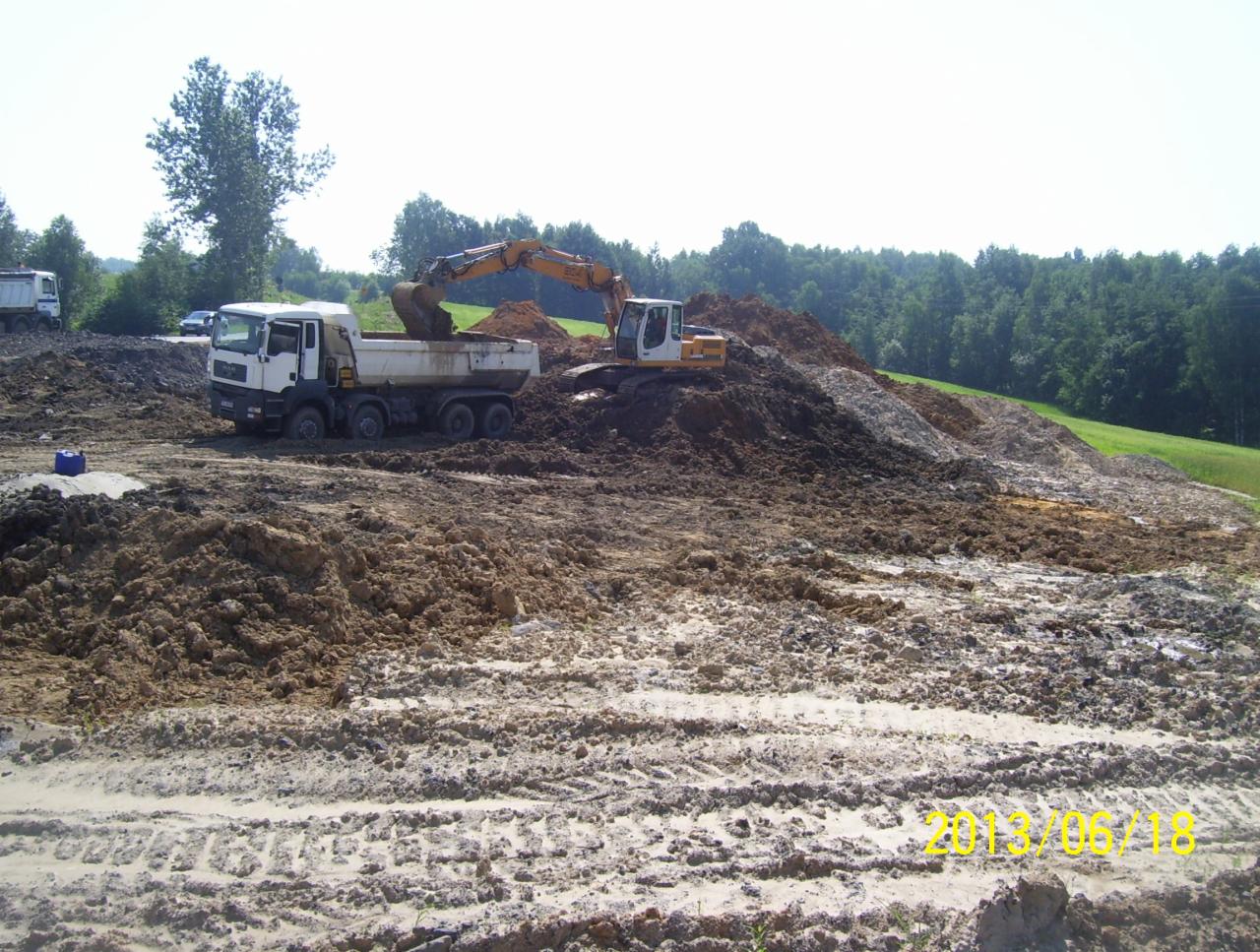 Żółta koparka ładuje ziemię na białą ciężarówkę MAN na placu budowy. W tle drzewa i niebo. Na pierwszym planie ślady opon na błotnistym terenie.